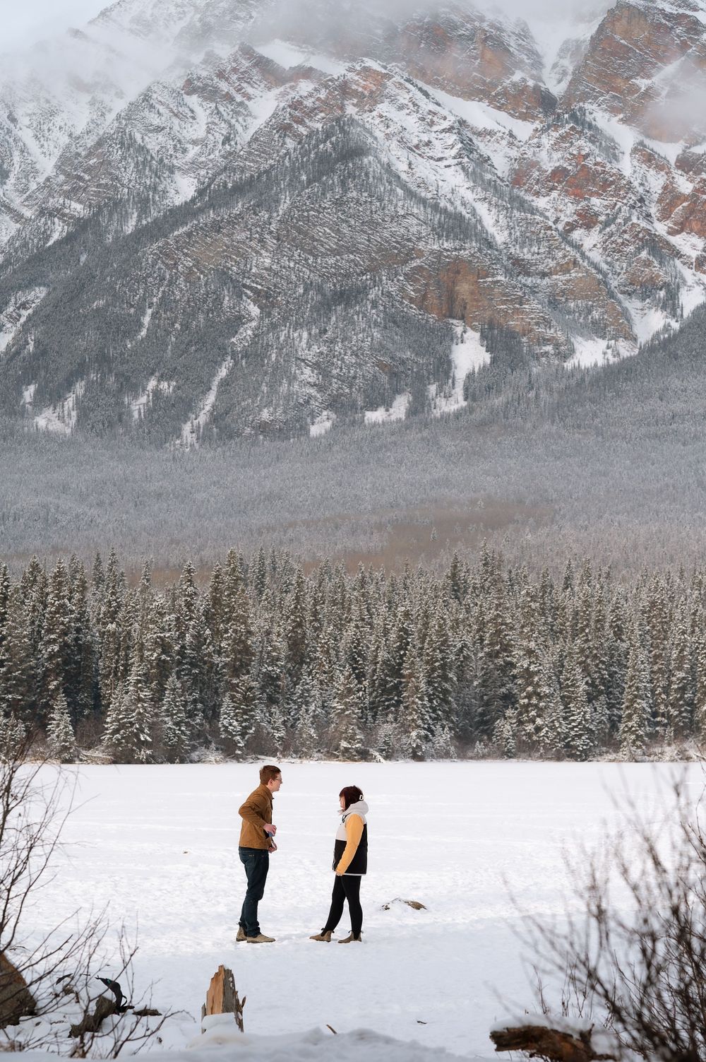 Two figures stand in deep snow with towering snow-capped mountains and pine forest in the background.
