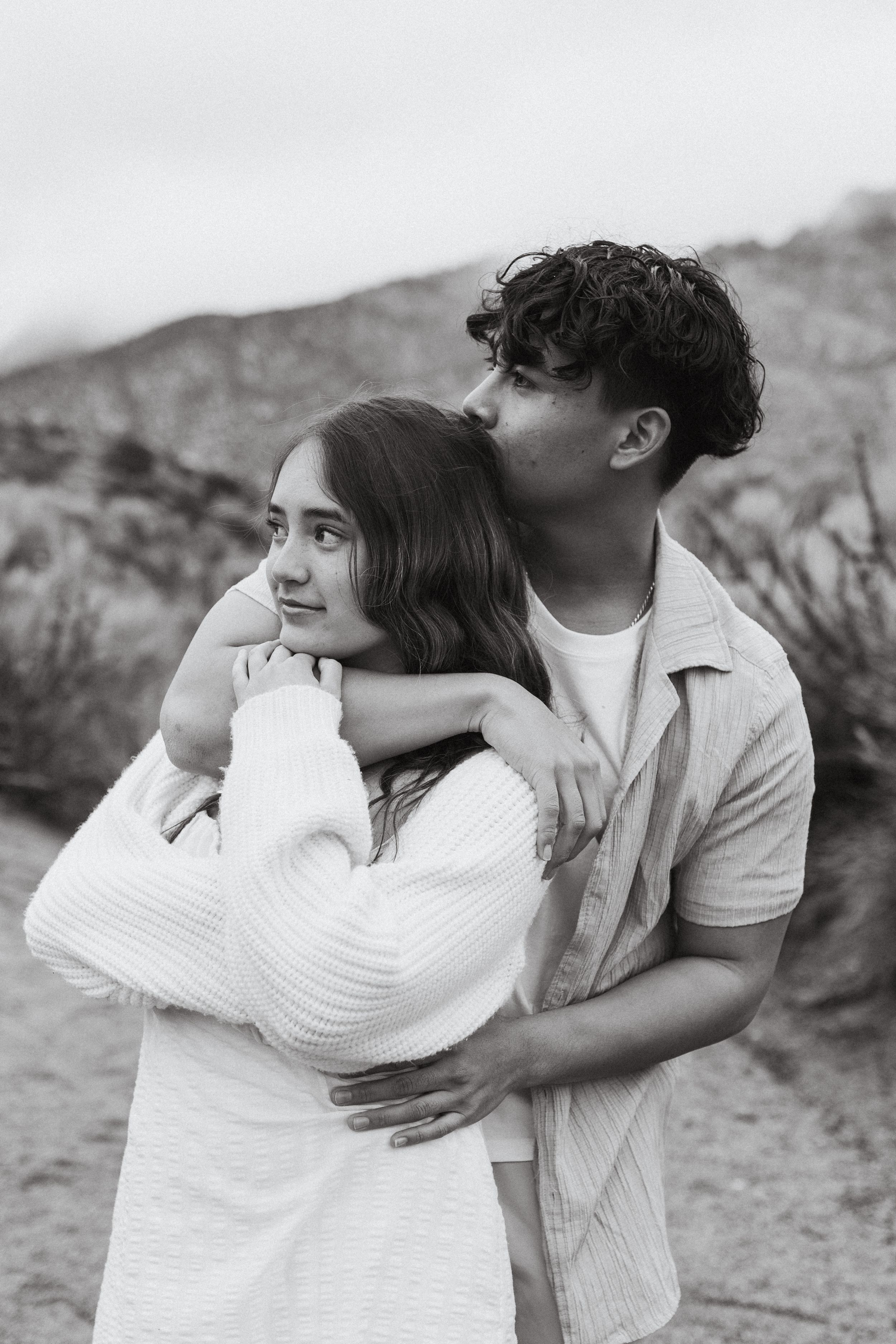 Black and white portrait of a couple embracing outdoors with mountains in the background.