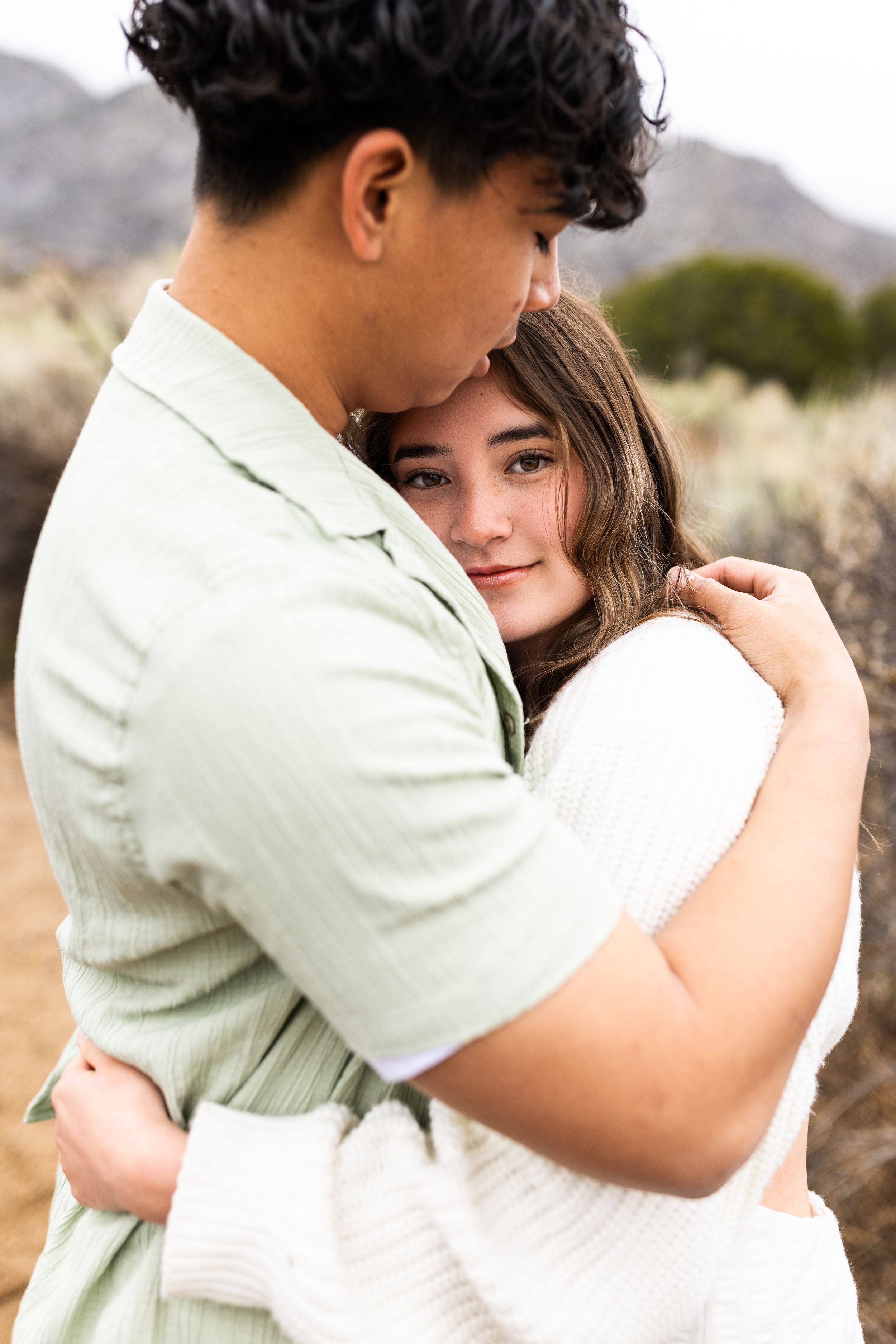 Romantic couple embraces outdoors with mountains and desert brush in the background on a sunny day.