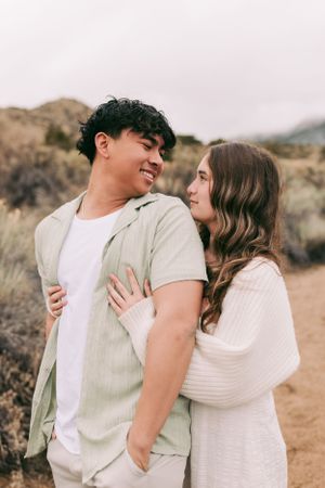 A couple in light colored clothing share an intimate moment while standing on a desert trail against mountainous backdrop.