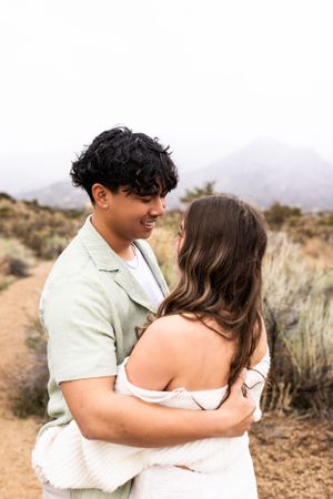 Romantic couple embracing in desert landscape with desert brush and mountains in background.
