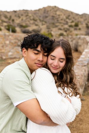 Couple shares intimate moment while hugging against desert mountain landscape in casual attire.