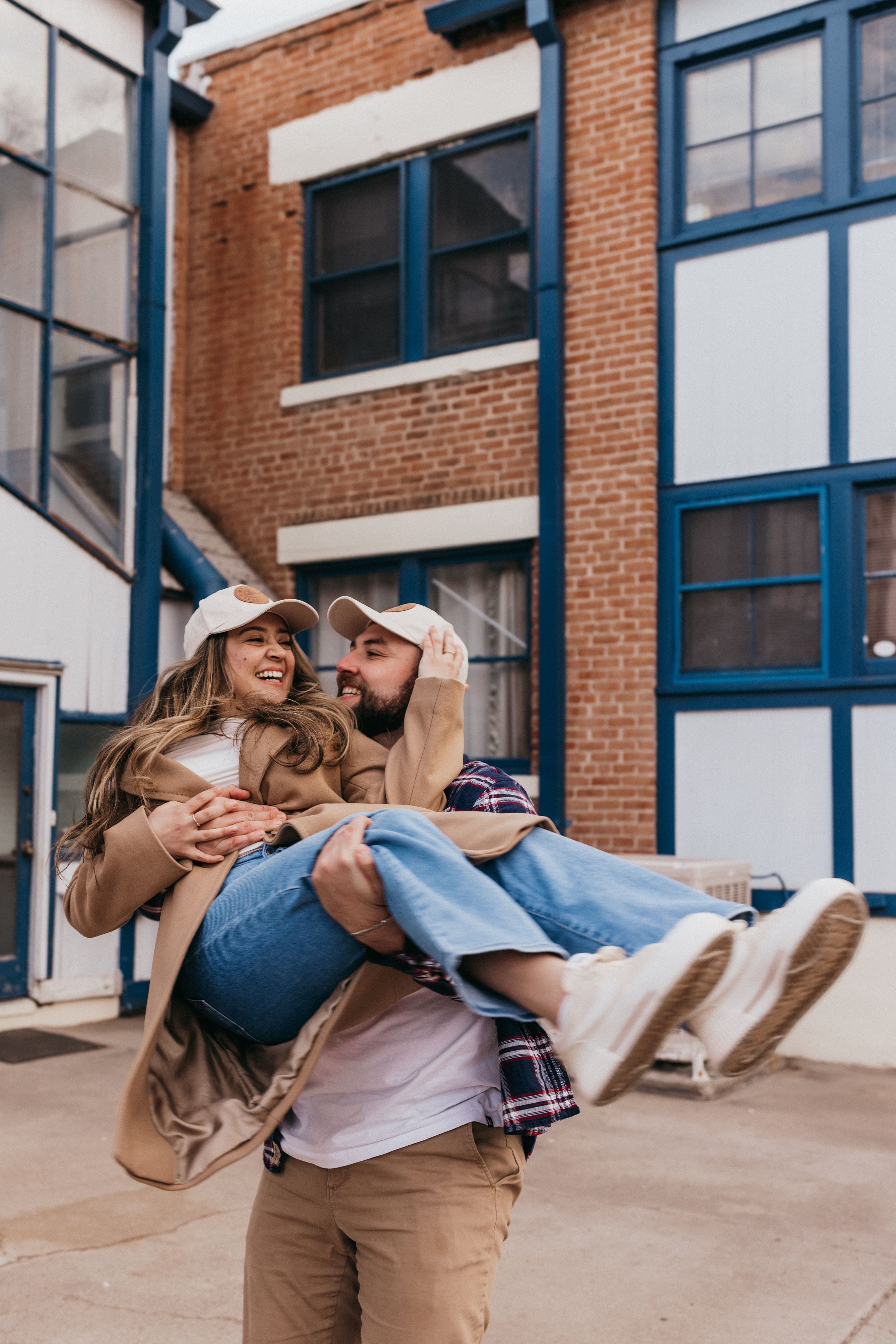 Two people share a joyful moment as one carries the other playfully in front of a brick building with blue trim.