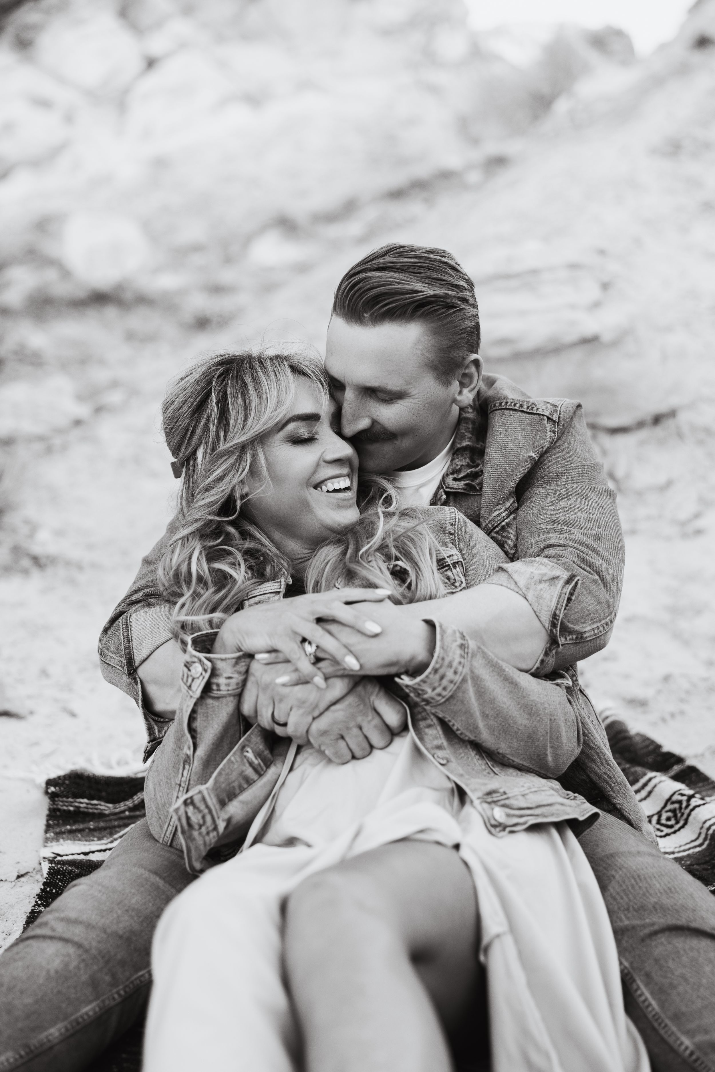 A romantic black and white portrait shows a couple in casual denim jackets embracing and laughing while sitting outdoors.