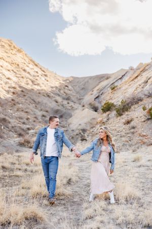 Couple walking hand in hand through scenic mountain terrain wearing matching denim jackets on sunny day.