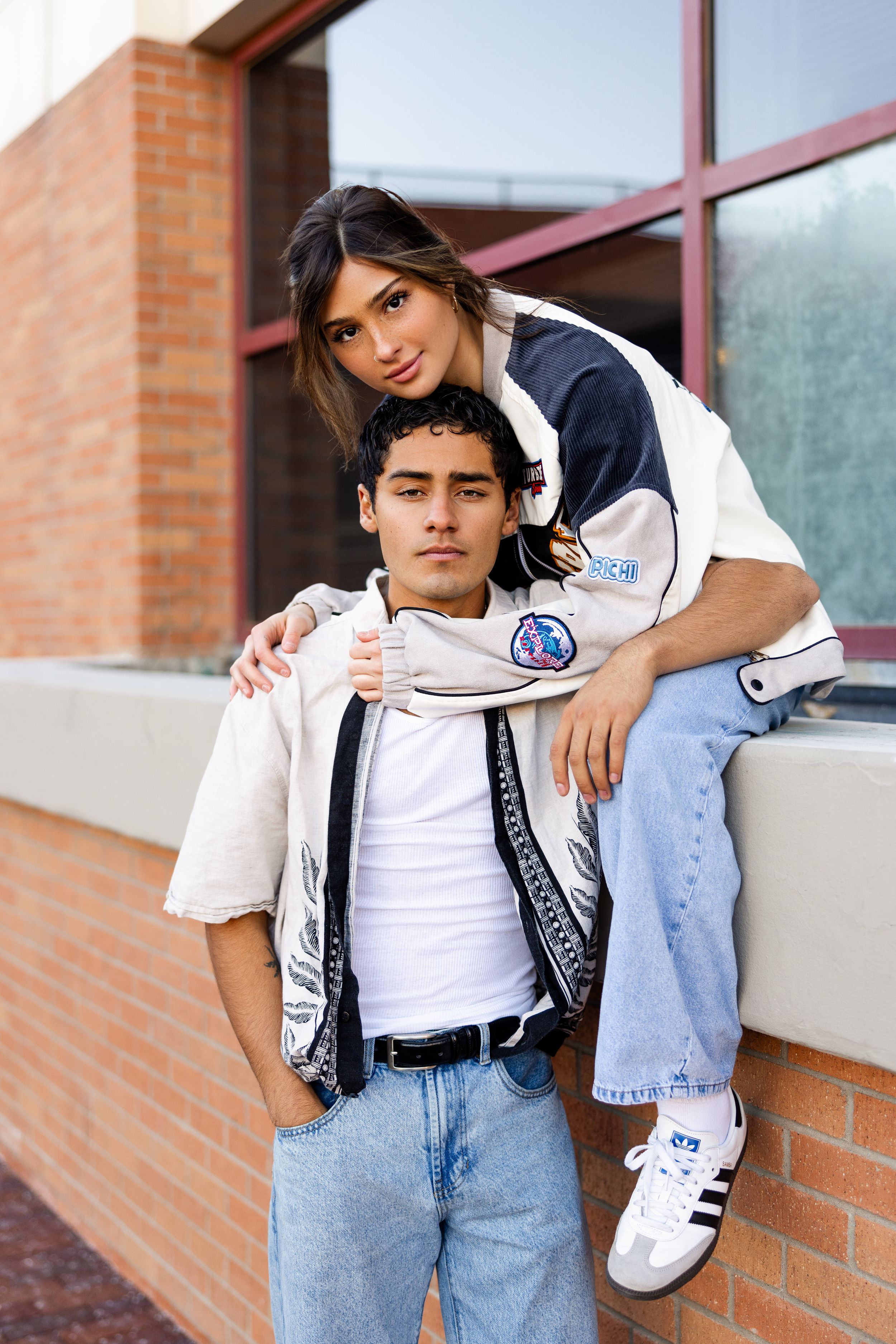 A young couple in casual sportswear poses against a brick wall, with one person sitting on ledge and the other standing.
