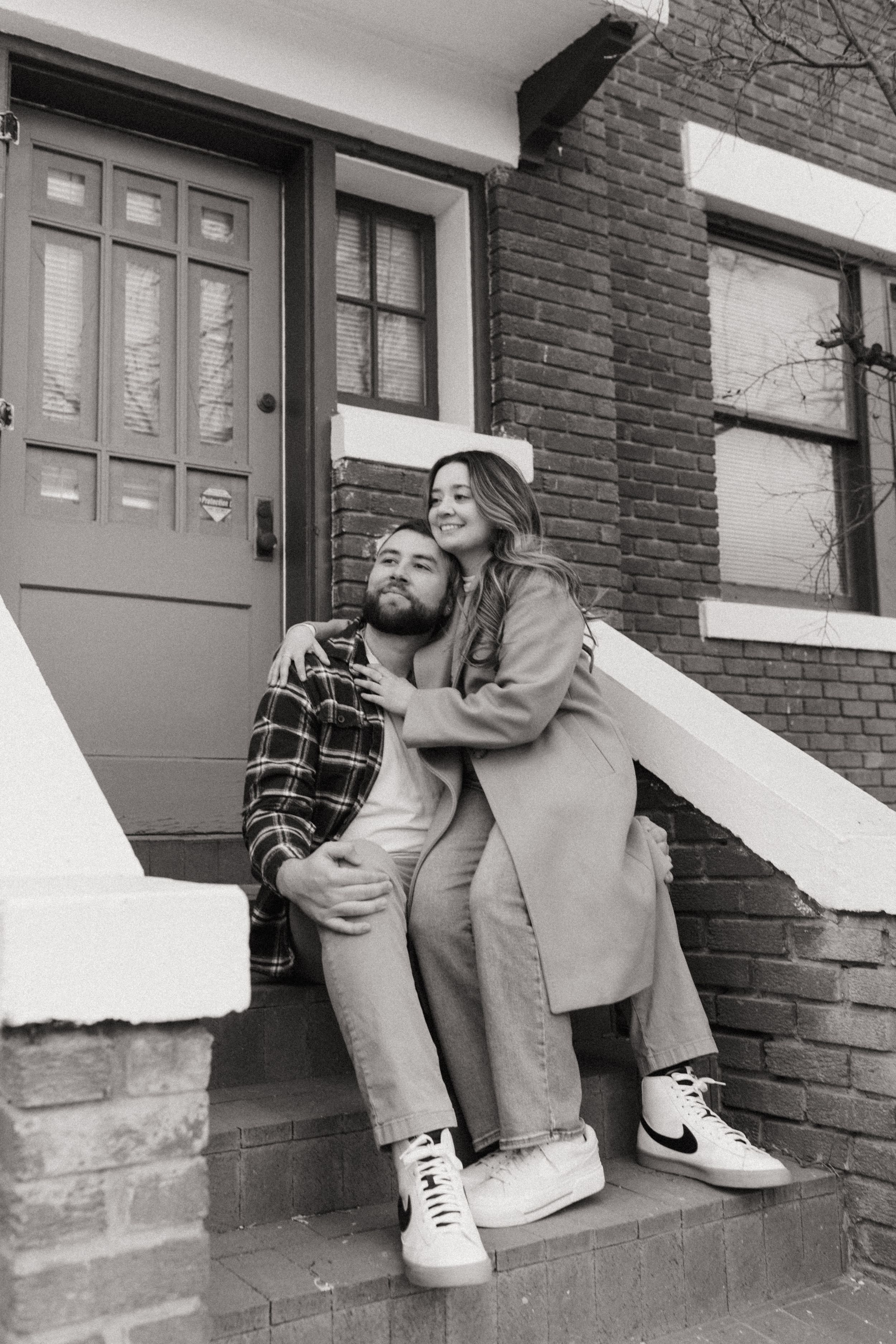 A black and white photo of a stylish couple sitting on steps outside a brick building with a vintage aesthetic.