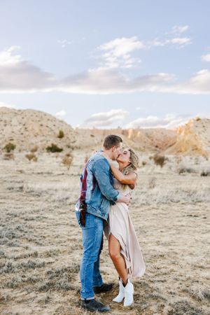 A couple embraces in a scenic desert landscape at sunset with mountains and soft clouds in the background.