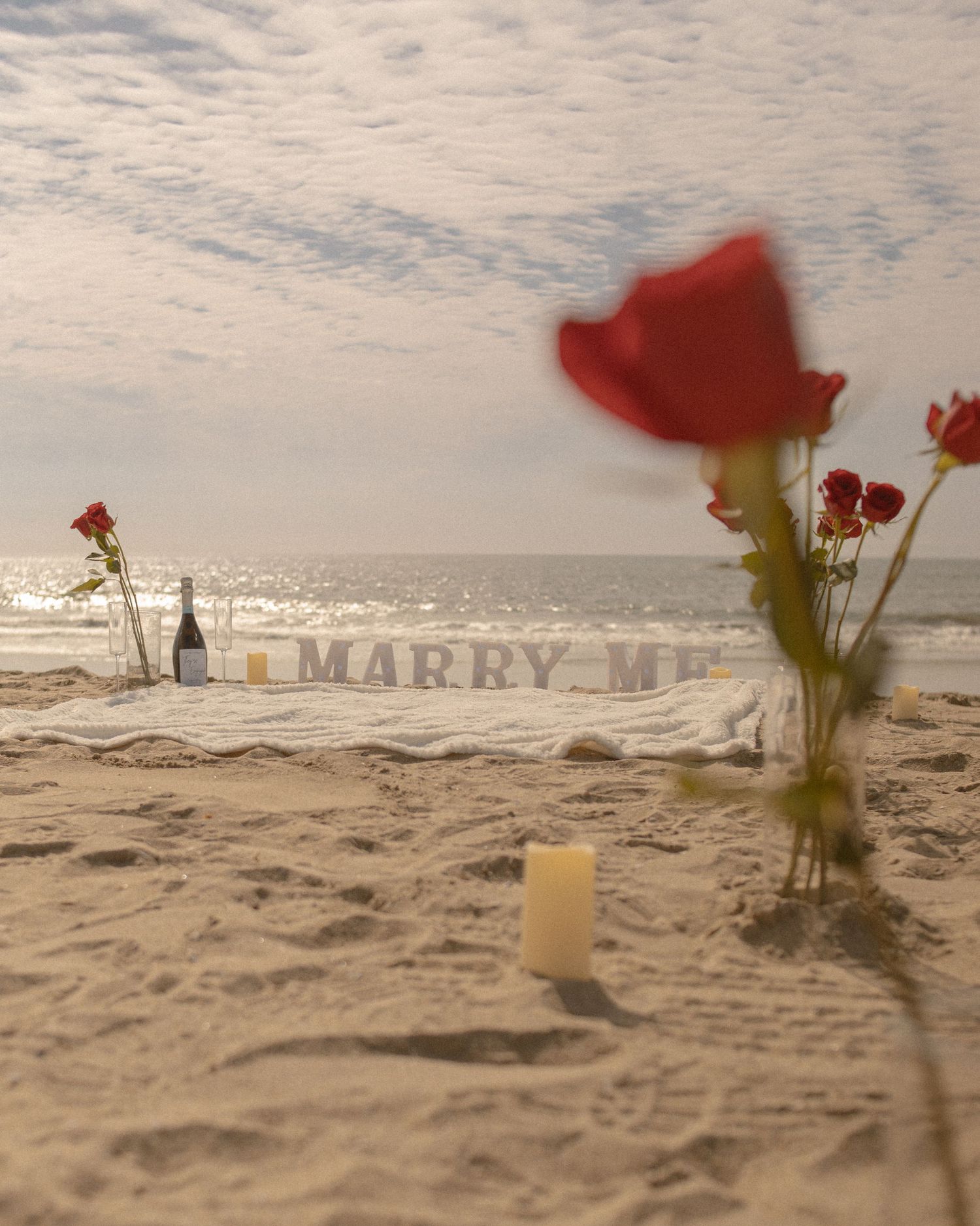 Romantic beach proposal setup with "MARRY ME" letters, red roses, and candles at sunset on the sandy shore