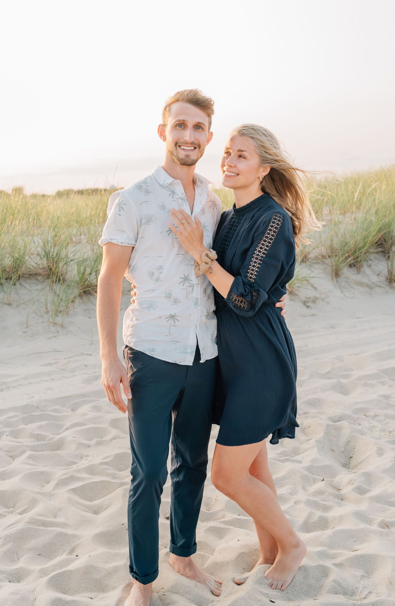 A young couple stand in front of the dunes on the beach in spring lake nj.