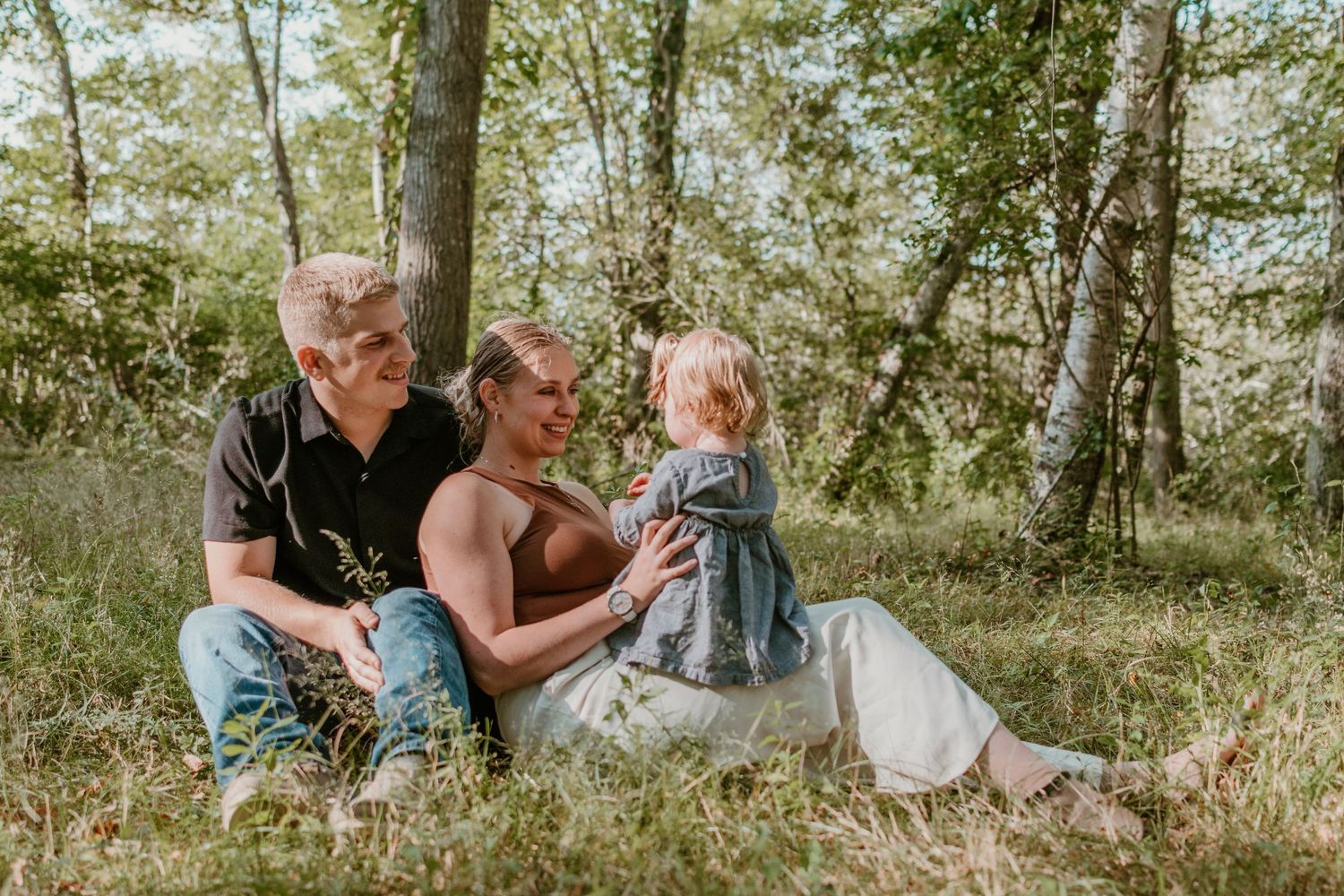 People spending time together outdoors sitting in a sunny wooded area during a casual family photo session.