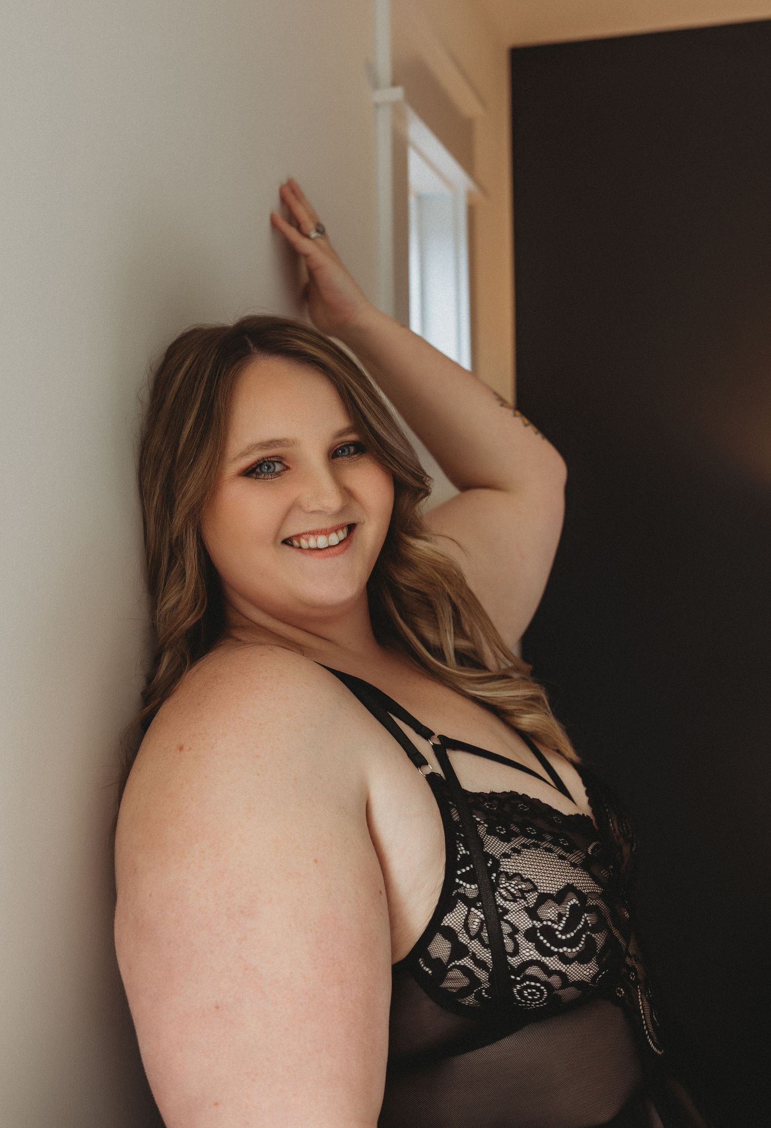 Boudoir portrait of person in lace lingerie leaning against wall with natural lighting.
