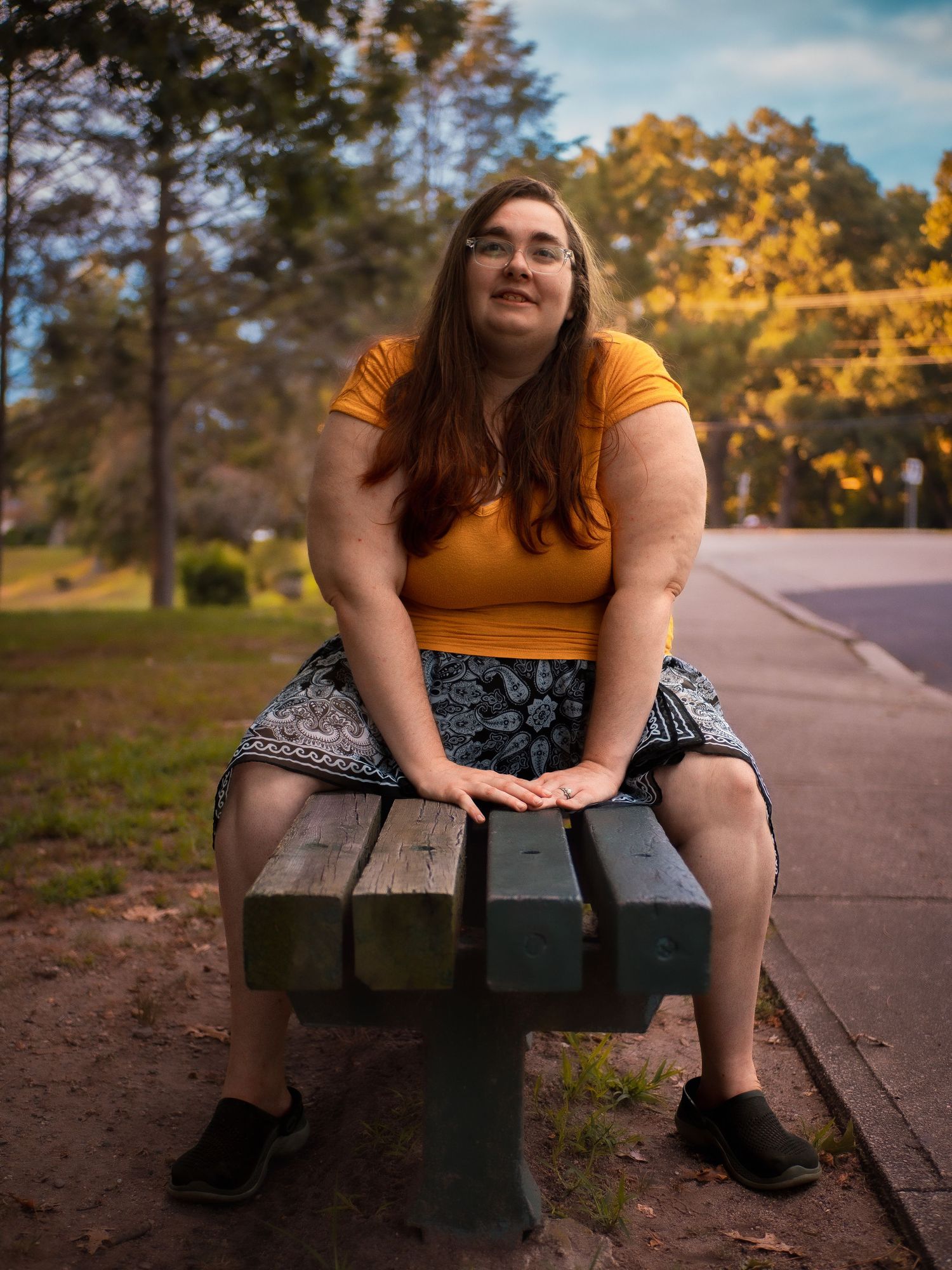 Person in yellow crop top sitting on park bench during golden hour with autumn trees in background.