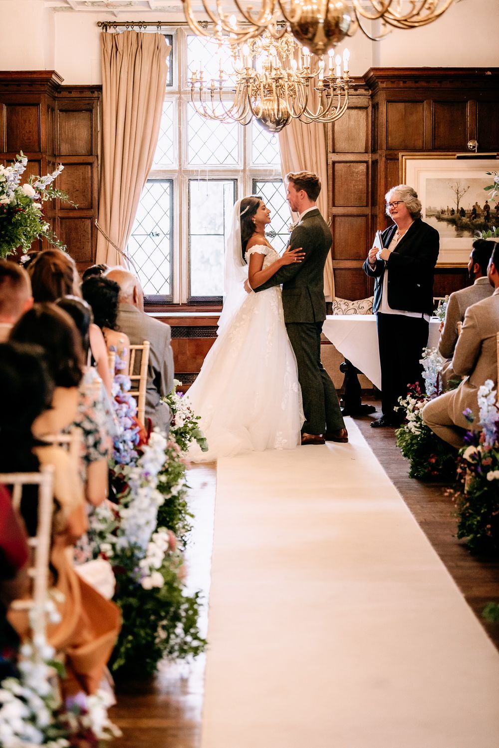 Wedding ceremony in progress inside an elegant wood-paneled room with chandeliers and floral decorations.