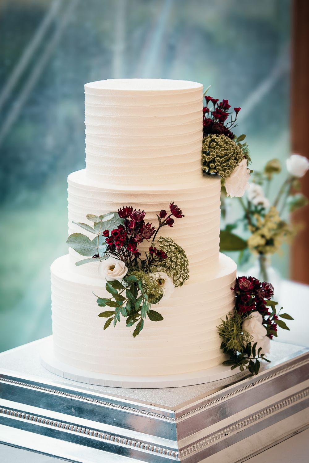 White wedding cake decorated with burgundy flowers and greenery on a silver stand.