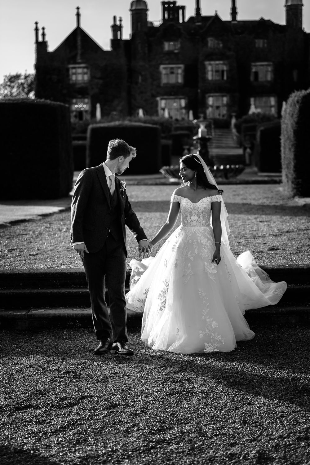 Elegant wedding couple walking across a grassy lawn with historic buildings in the background.