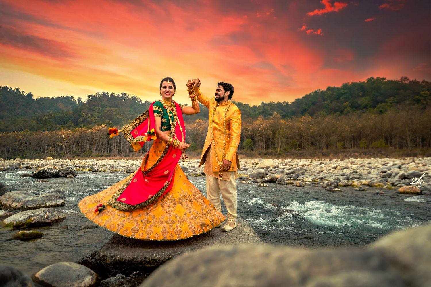 Couple in traditional Indian attire dance together on rocky riverbank during dramatic orange sunset in mountains.
