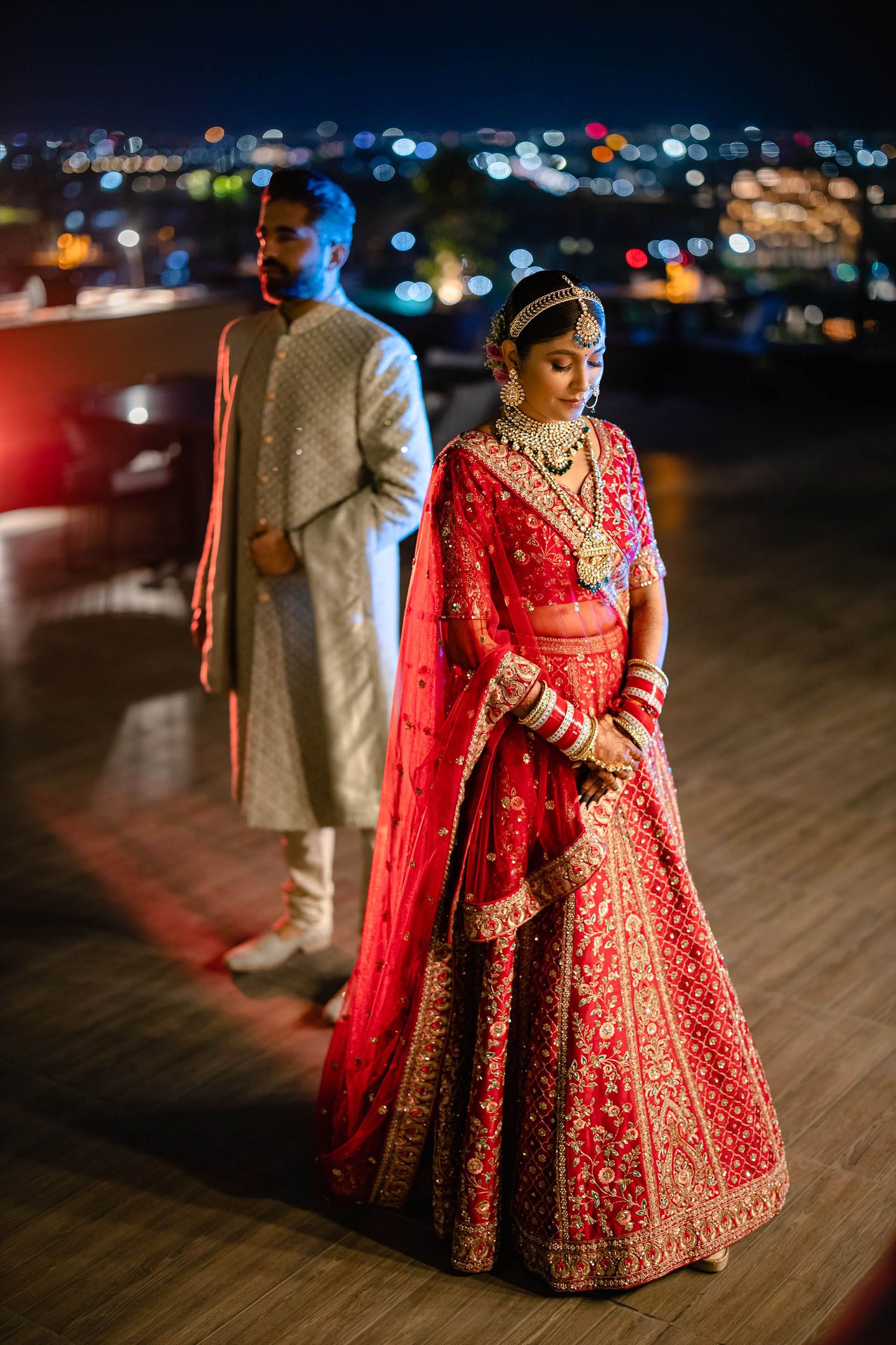 Traditional Indian wedding couple in evening attire standing on rooftop with city lights in background at night.