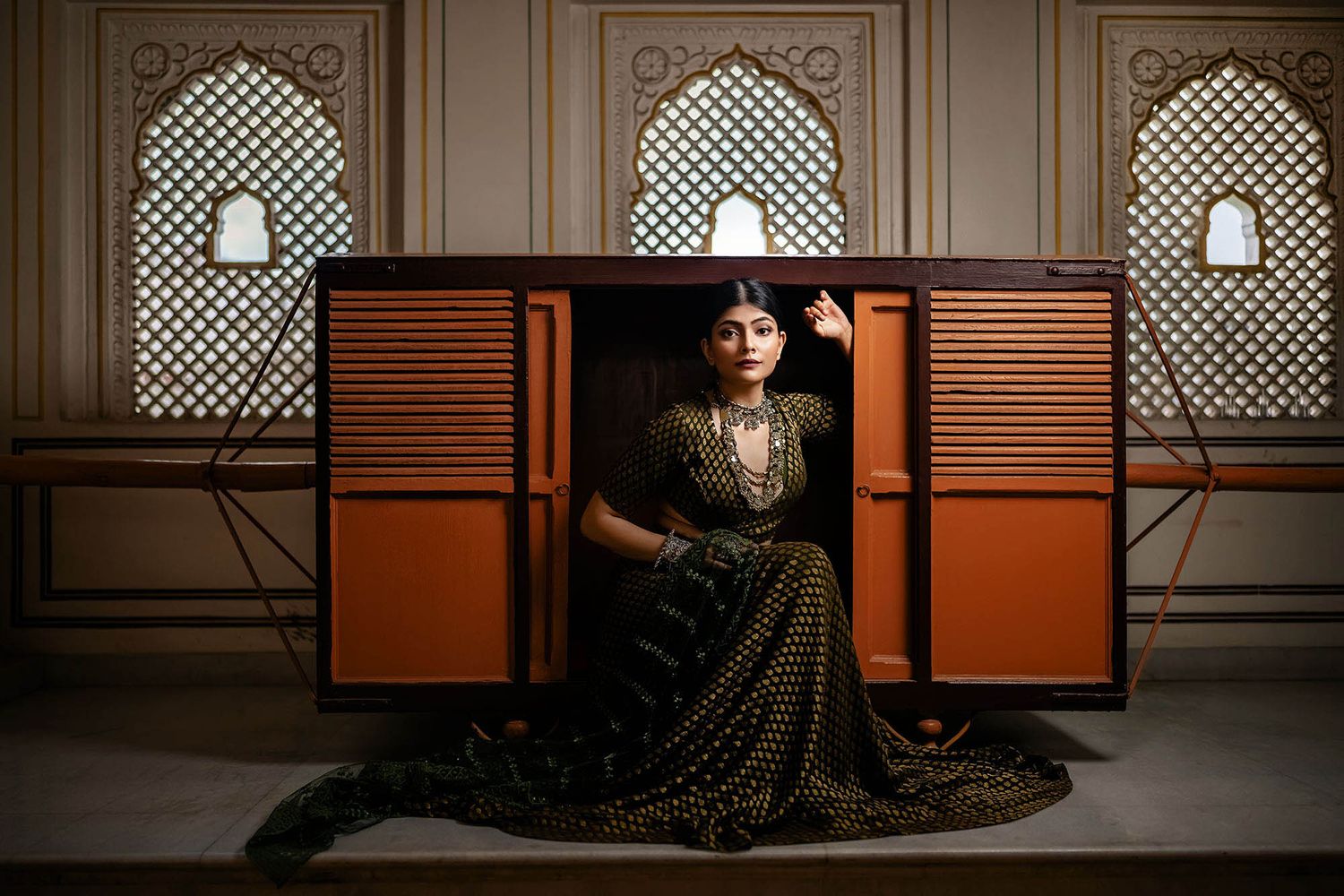 Elegant model in ornate green lehenga poses against vintage orange shutters at luxury Indian palace interior.