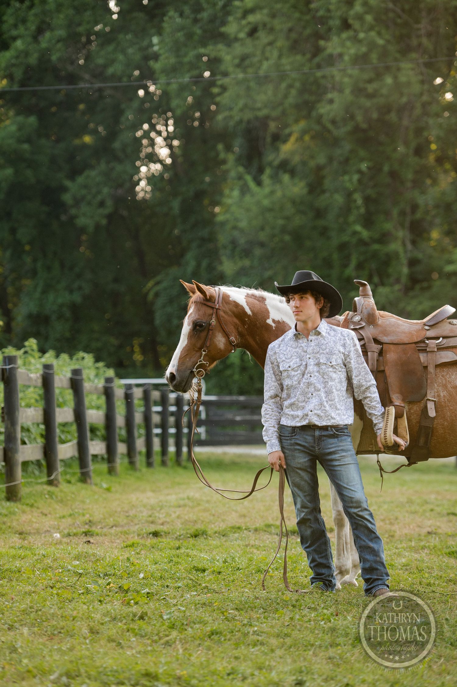 A person in western wear leads a paint horse along a wooden fence line at sunset.
