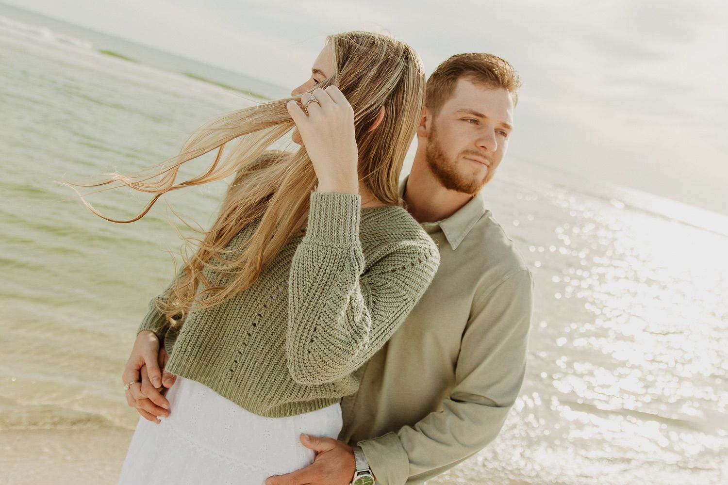 A couple embraces on a sunny beach while her long blonde hair flows in the wind.