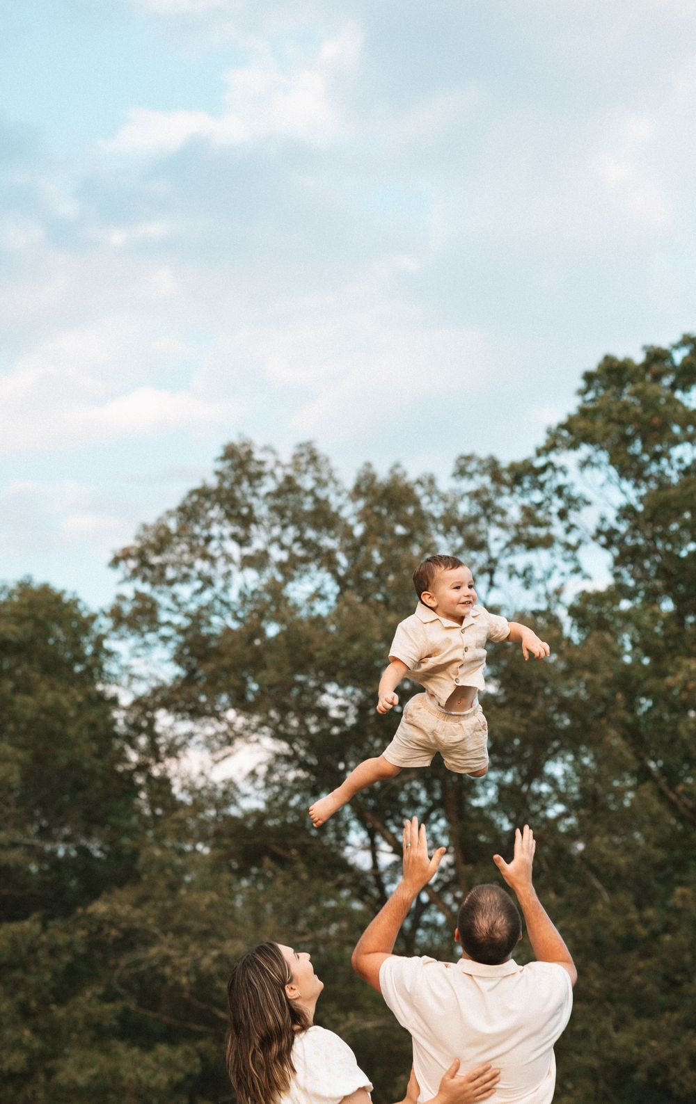 Joyful family playing and lifting child in the air against a backdrop of trees.