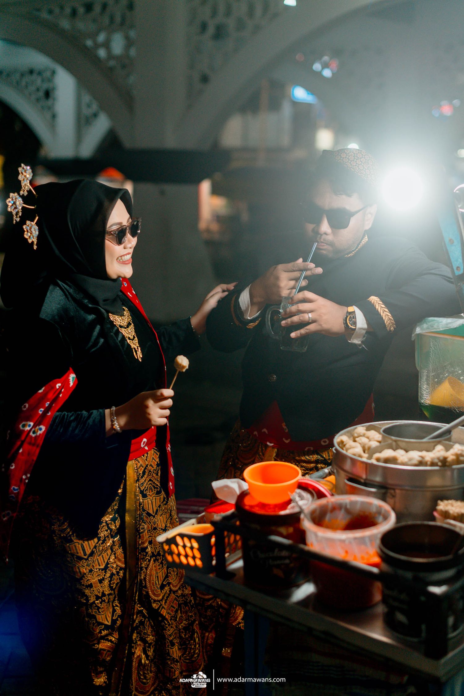 Guests in traditional attire gather around a dining setup with drinks and food at an evening celebration.