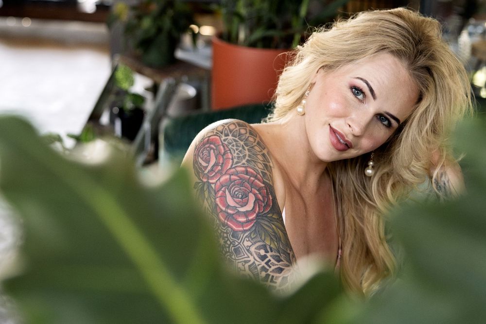 A blonde woman with a colorful shoulder tattoo smiles warmly while posing in a natural outdoor setting.
