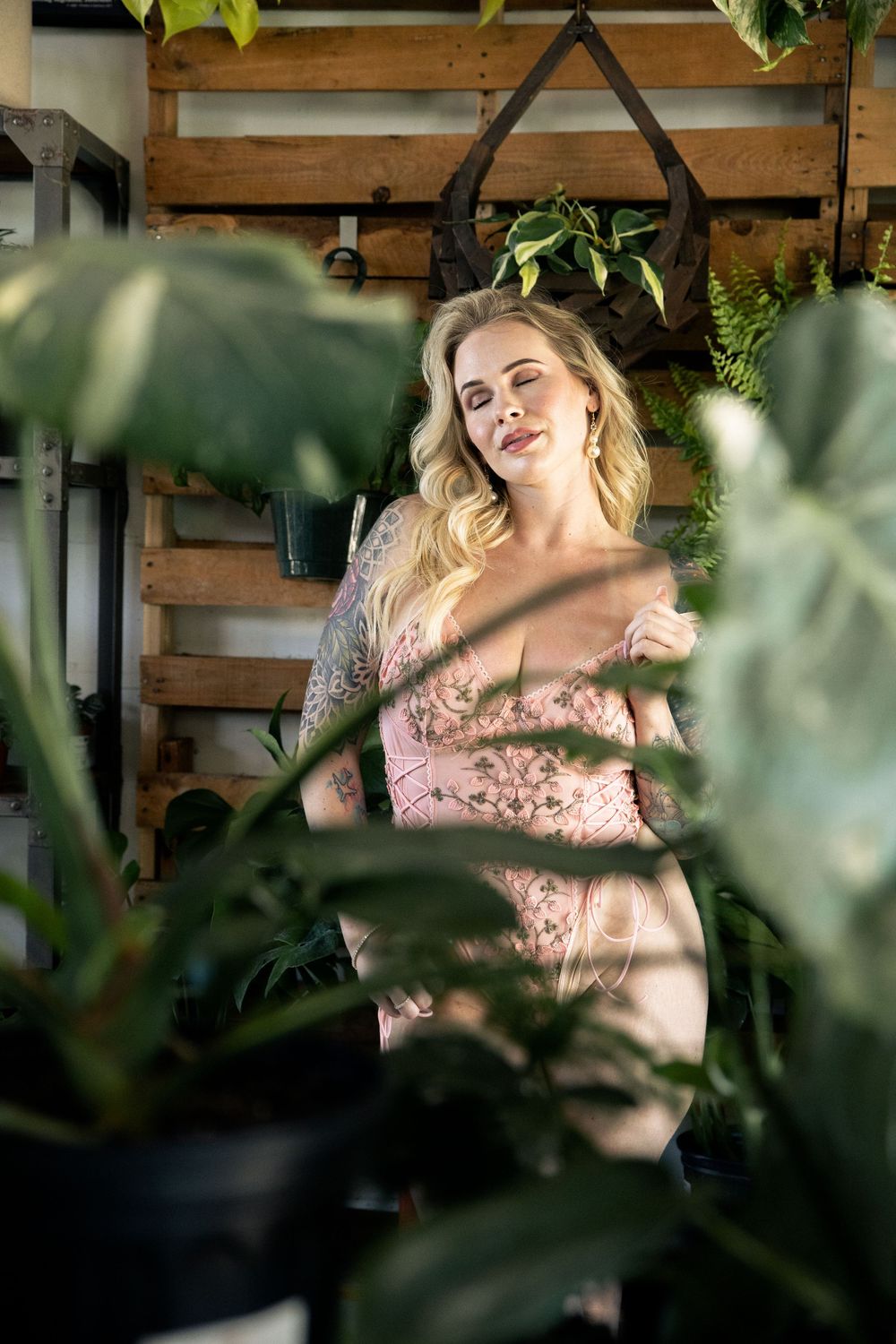 Pink floral dress seen through lush green plants in a sunlit indoor garden setting with wooden wall details.