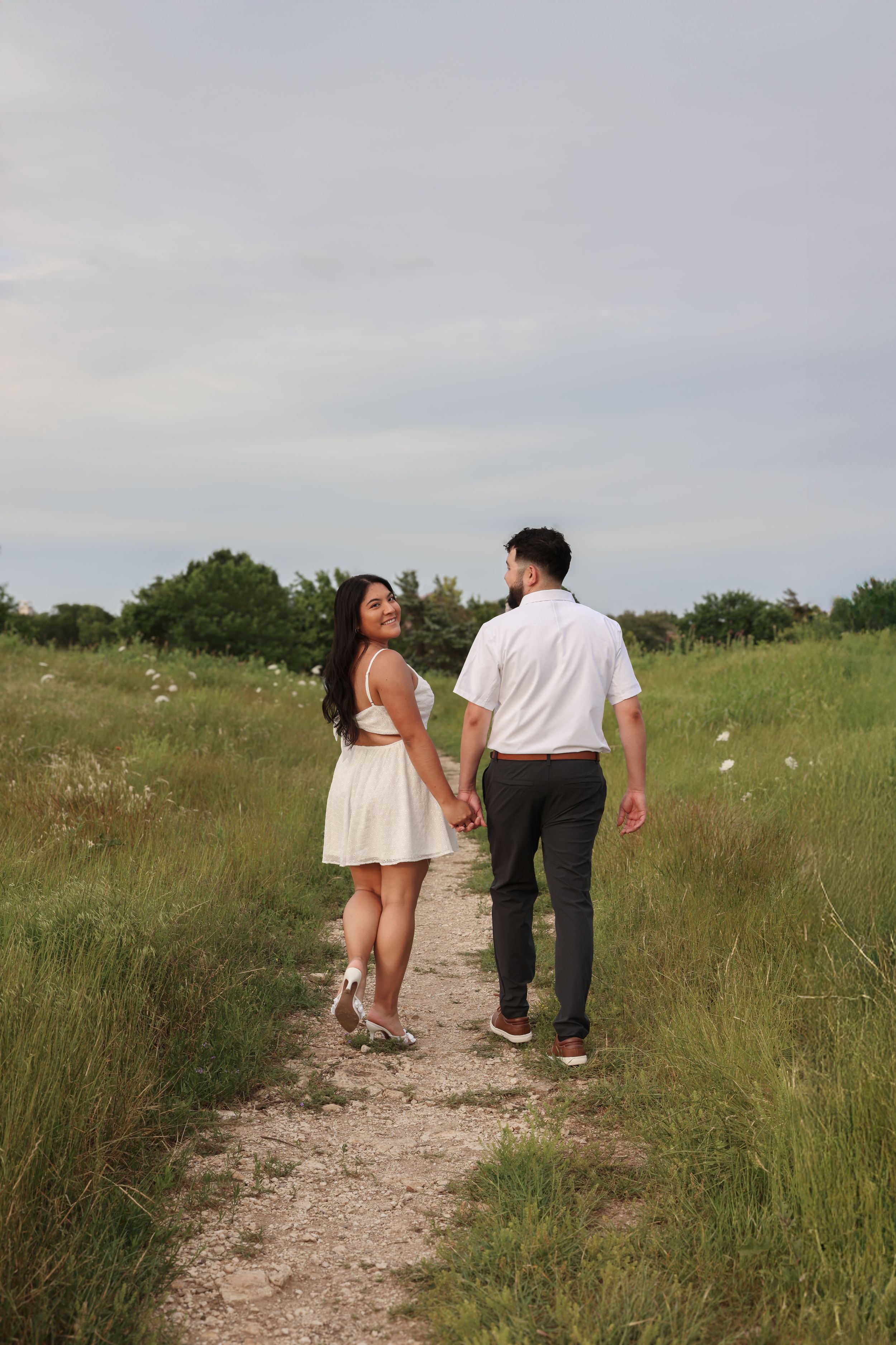 A couple walks along a grassy path on a cloudy day wearing casual outfits.