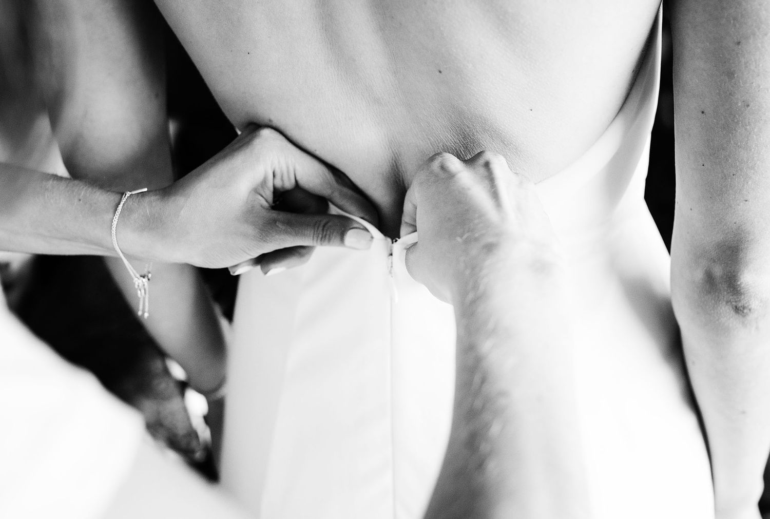 Close-up black and white photo of hands fastening the back of a white wedding dress.