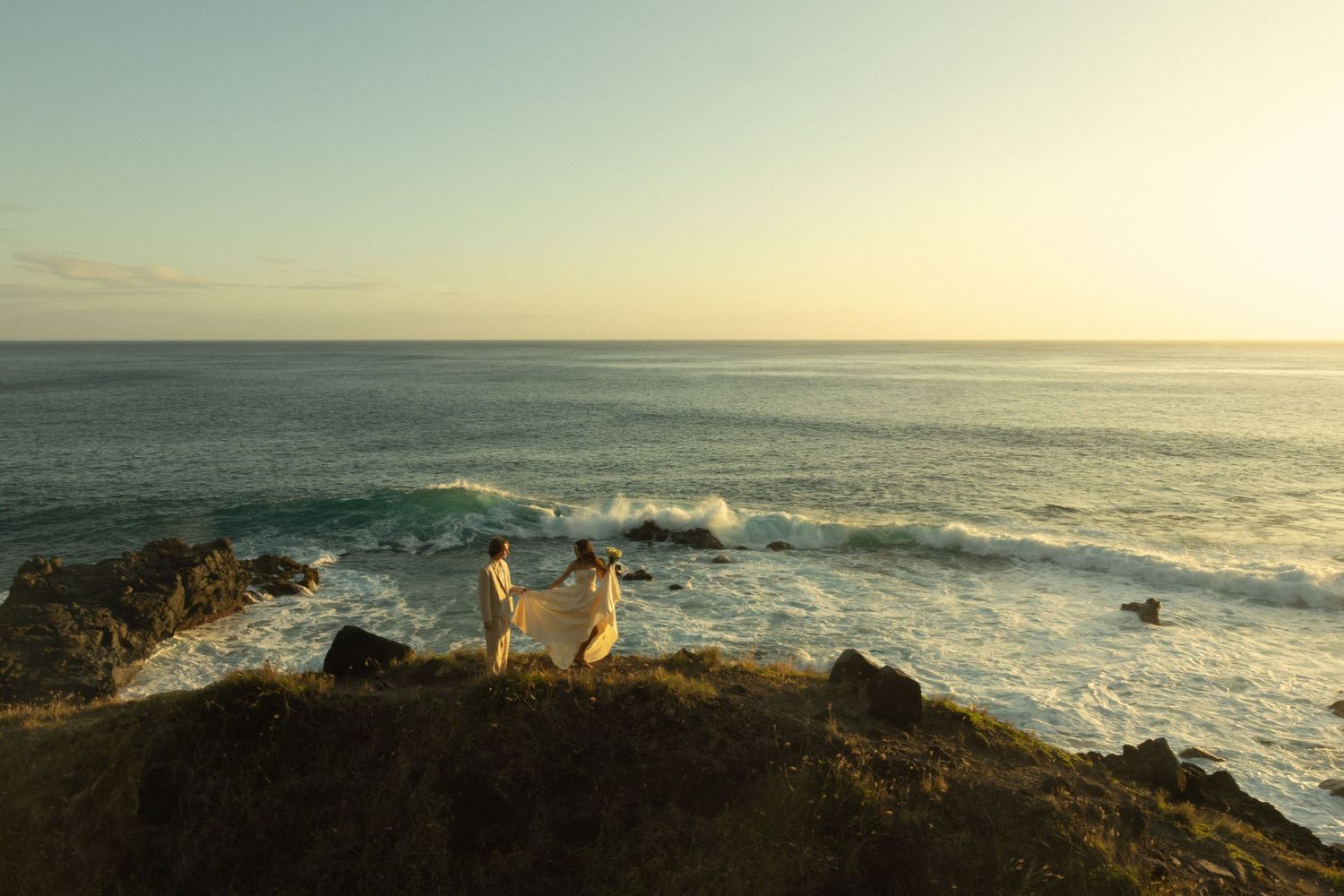 Two figures stand on rugged coastal cliffs at sunset overlooking a dramatic ocean landscape with crashing waves.