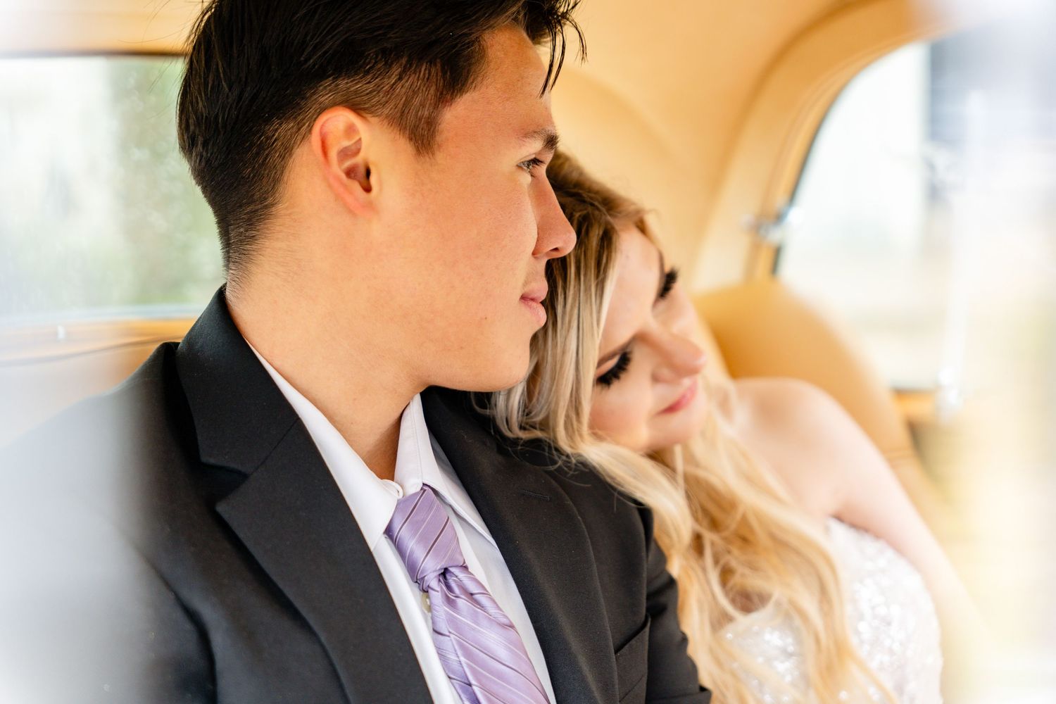 A couple rests together in a luxury car interior, with the person wearing a dark suit and purple tie.