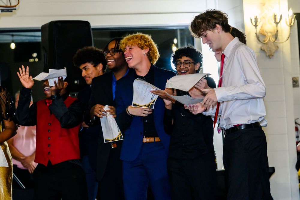 Young people in formal attire stand together reading from papers during what appears to be a performance or ceremony.