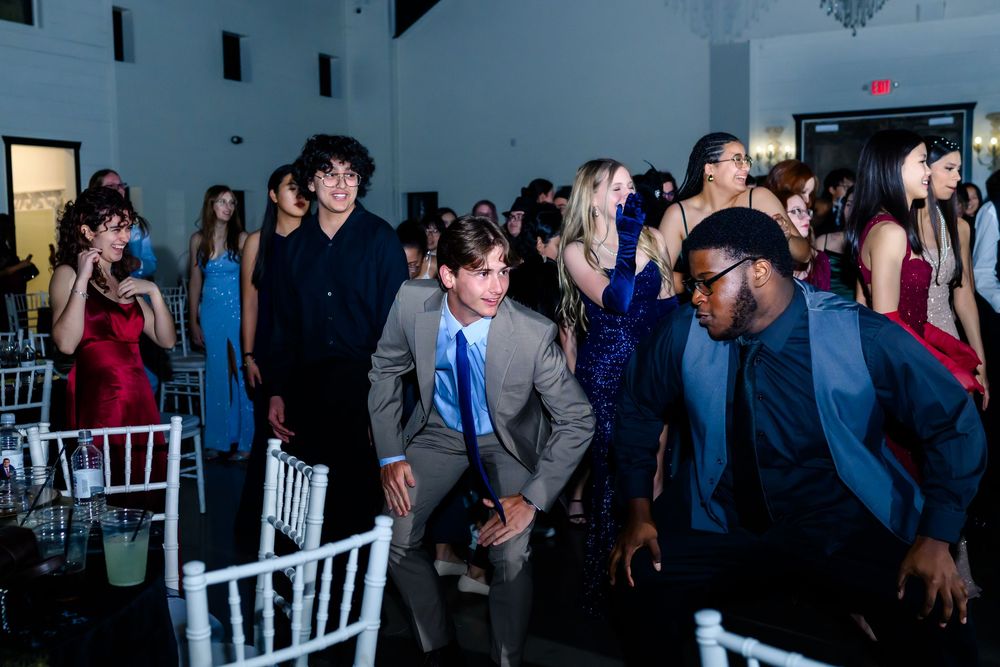 Wedding guests dance and celebrate on the dance floor at an indoor reception venue with white chairs.