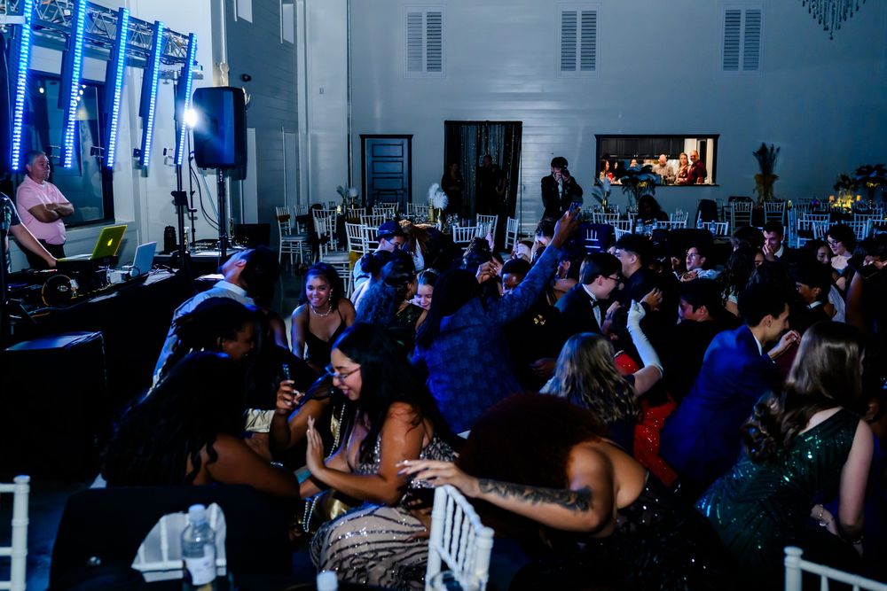 A crowd of people dancing at a party venue with blue lighting and white plastic chairs.