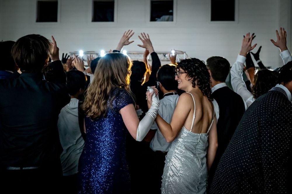 Party guests in evening attire dancing and raising their hands on a dimly lit dance floor at a formal celebration.