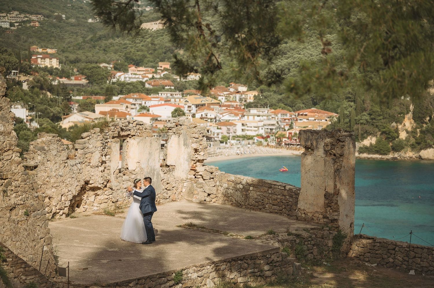 Scenic wedding photography at stone ruins above Mediterranean coastal town with turquoise bay.