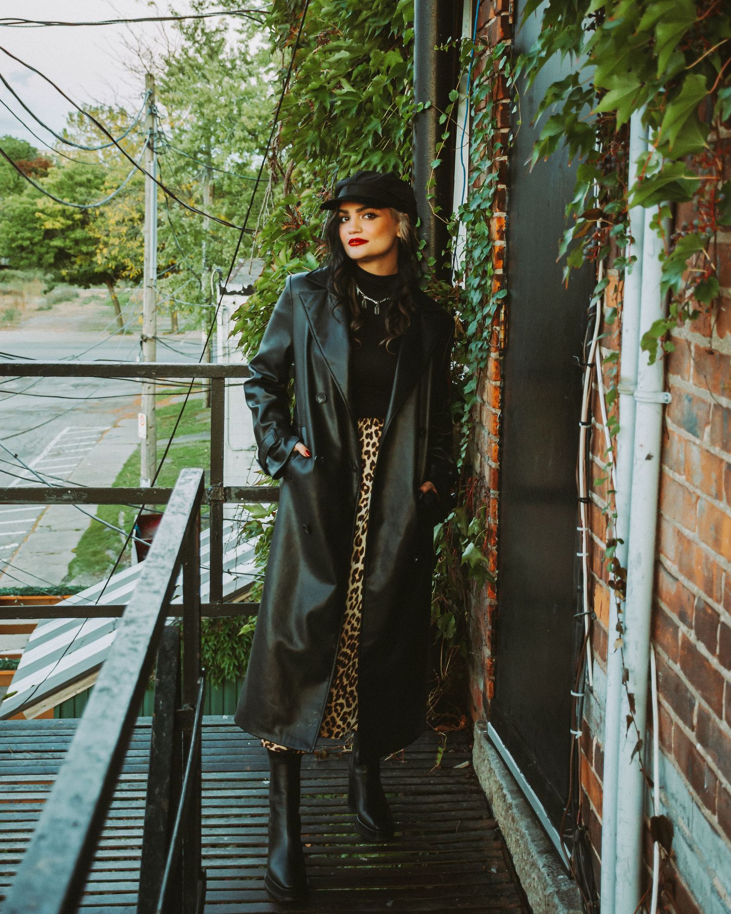 Moody fashion photoshoot on metal fire escape stairs with greenery and brick wall background.