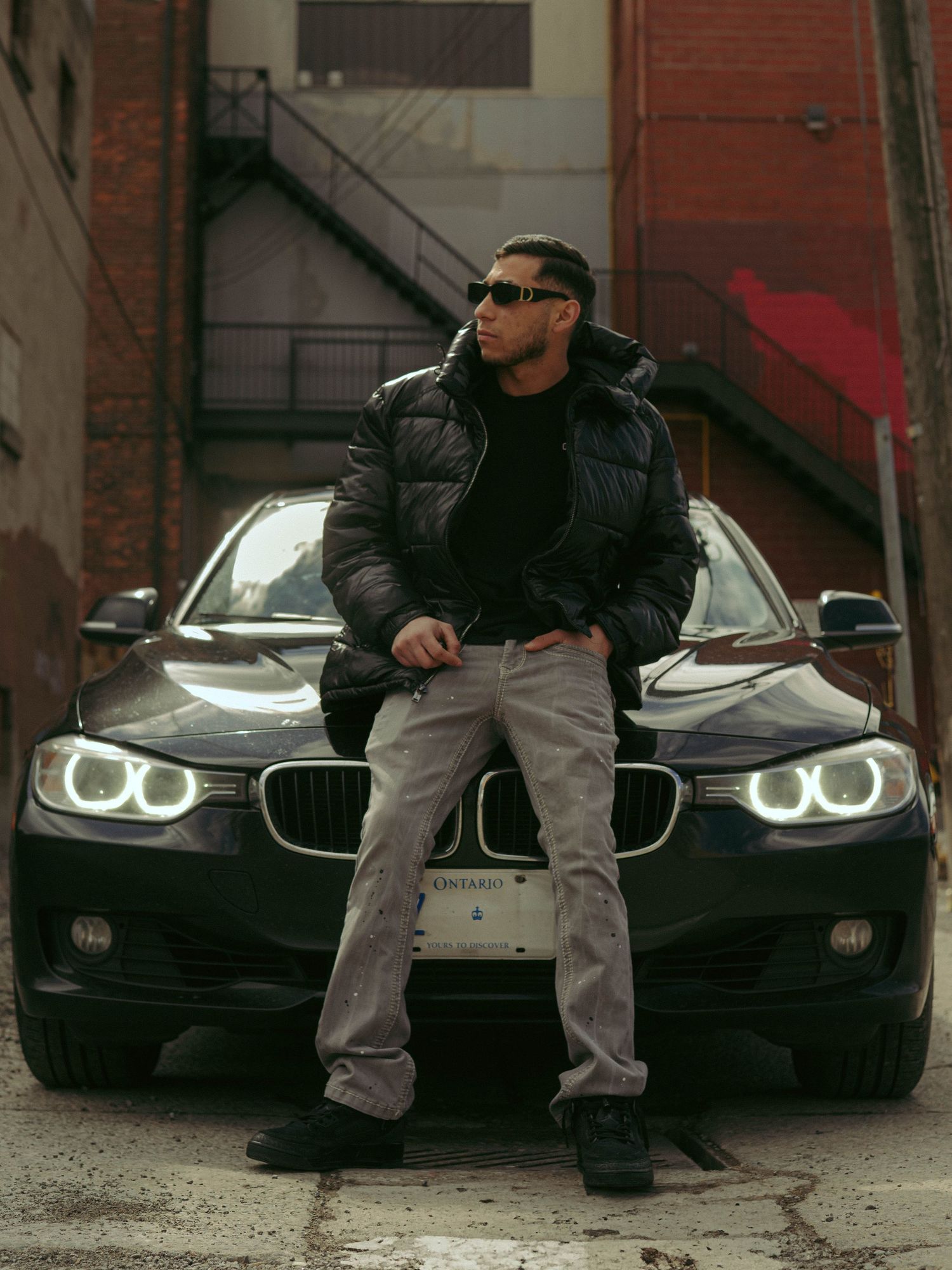 A series of photos showing someone posing with black luxury cars against a brick building backdrop.