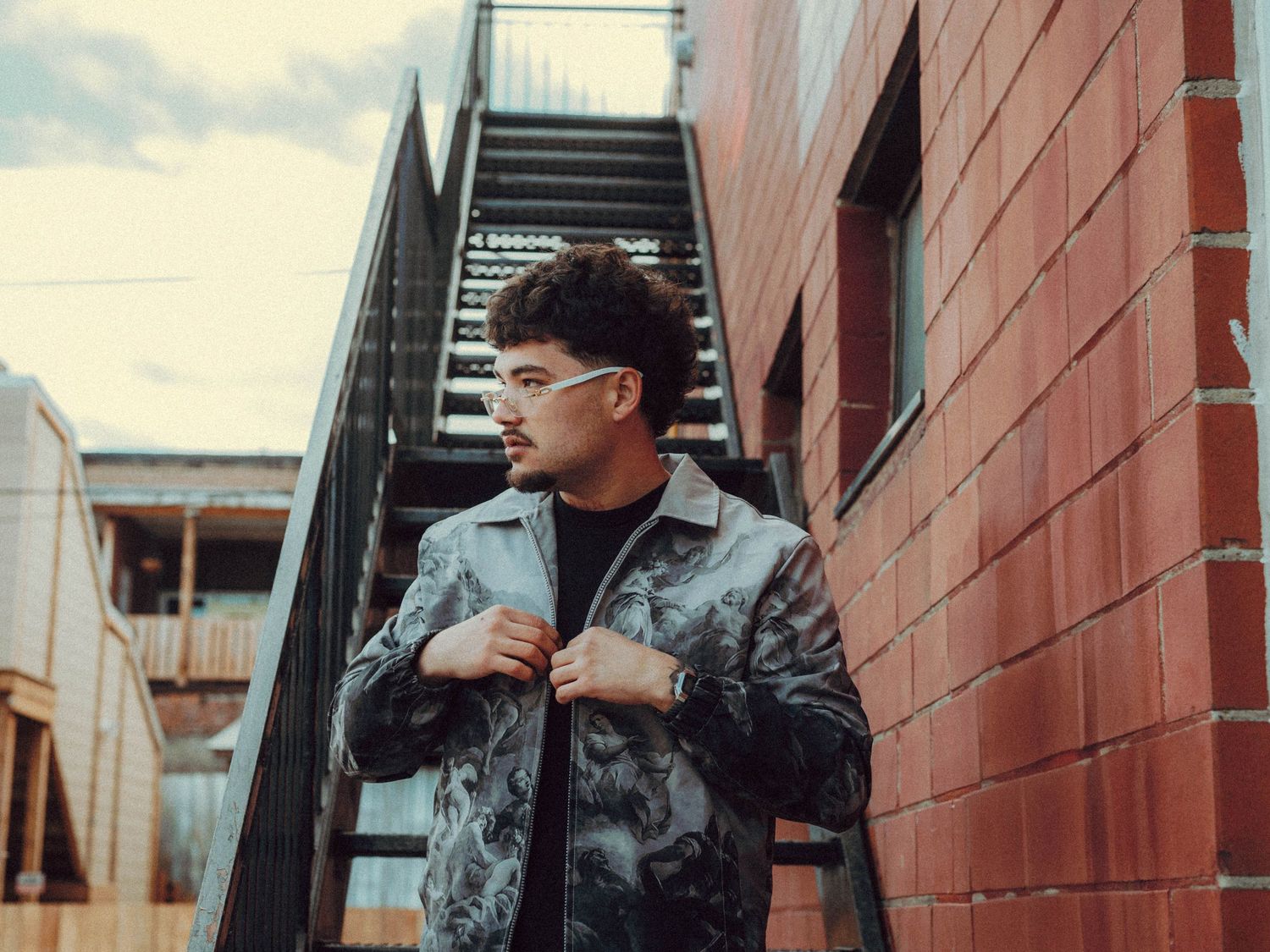 A person in casual wear poses on exterior stairs against a brick wall background.