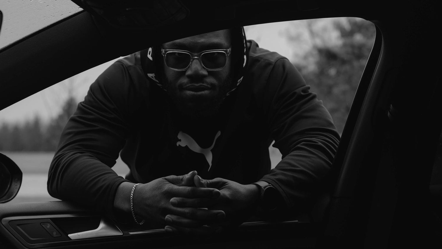 A person in sunglasses and dark clothing sits casually on a car in a moody black and white photo.
