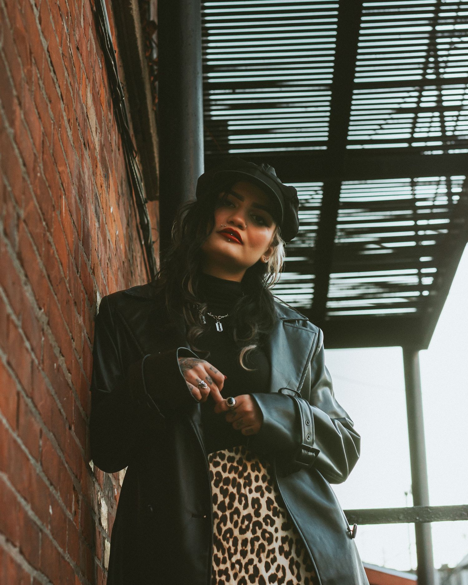 A figure in black clothing and leopard print stands against a brick wall with window blinds.