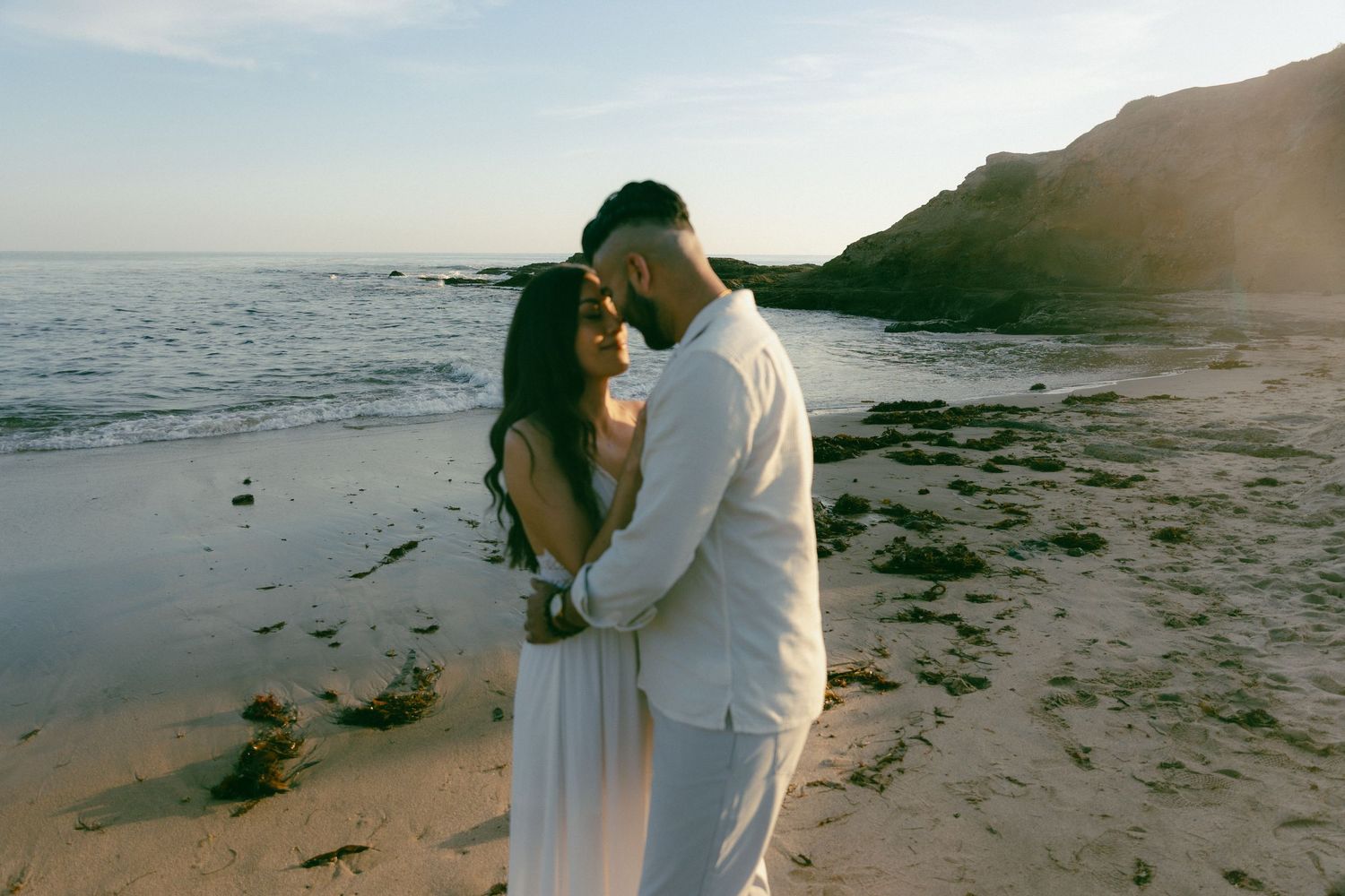 A couple in white outfits embrace during sunset on a scenic beach with rocky cliffs in the background.
