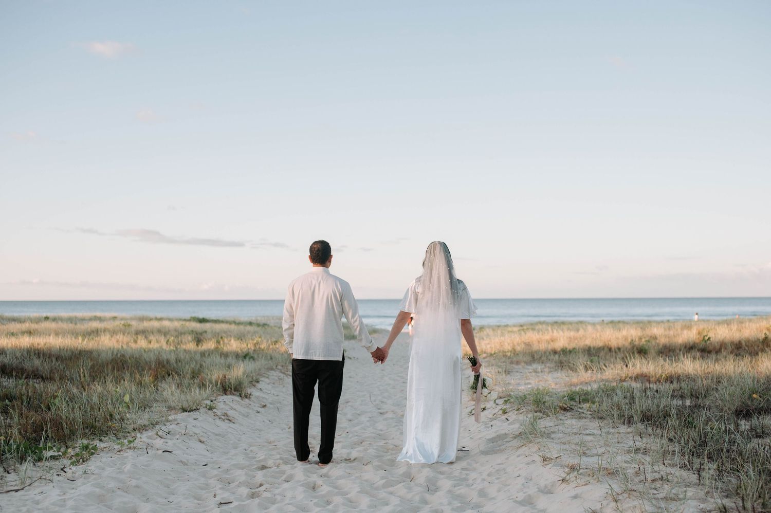 Couple walks hand in hand down dirt path towards ocean at sunset through coastal grassland.