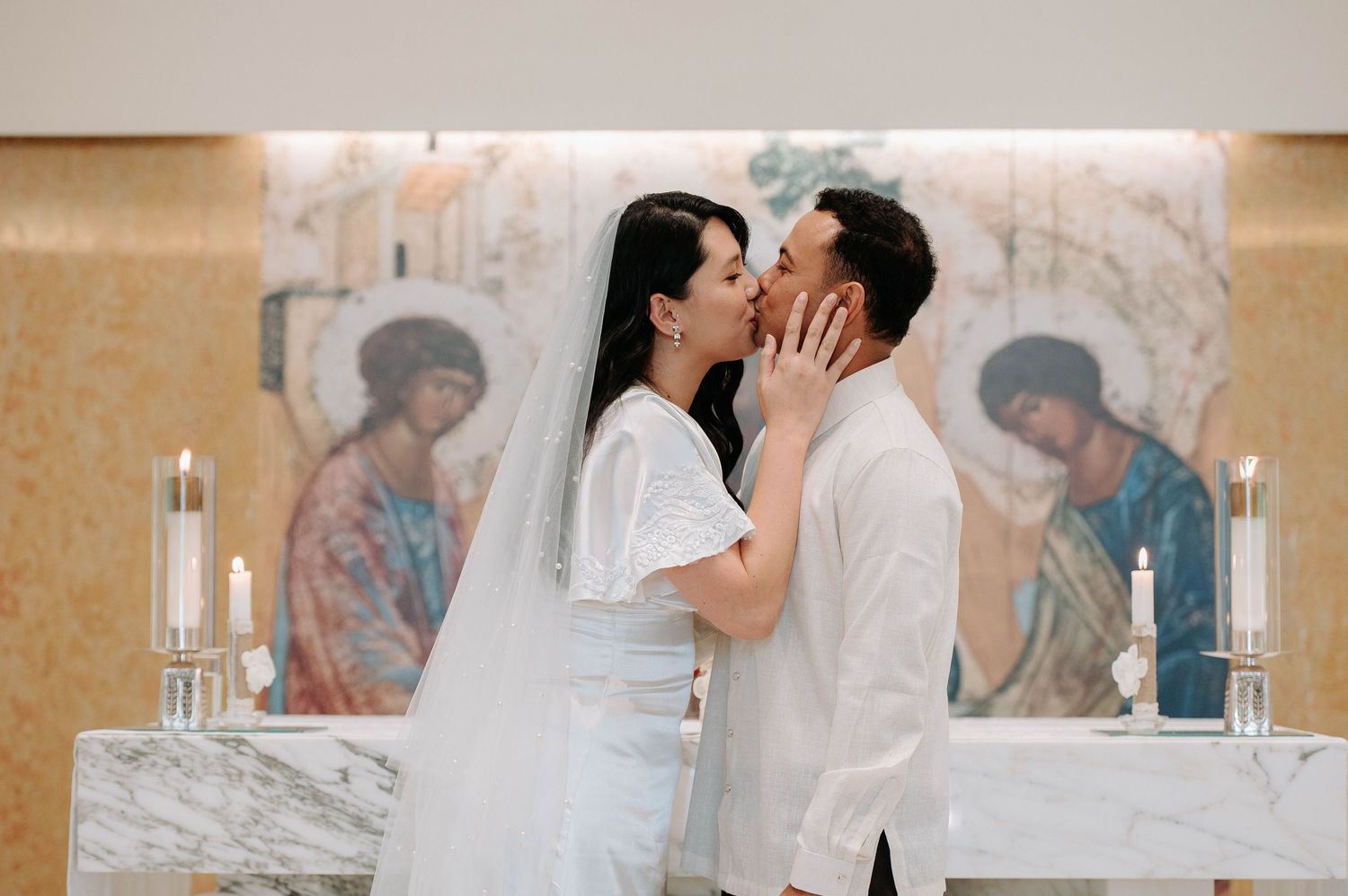 A tender wedding kiss shared at a church altar with religious artwork and marble walls in the background.