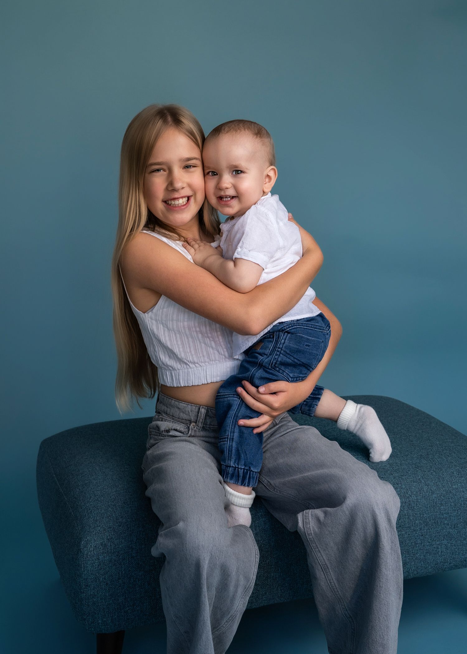 Young person holding a toddler while sitting together on a blue couch against a teal background.