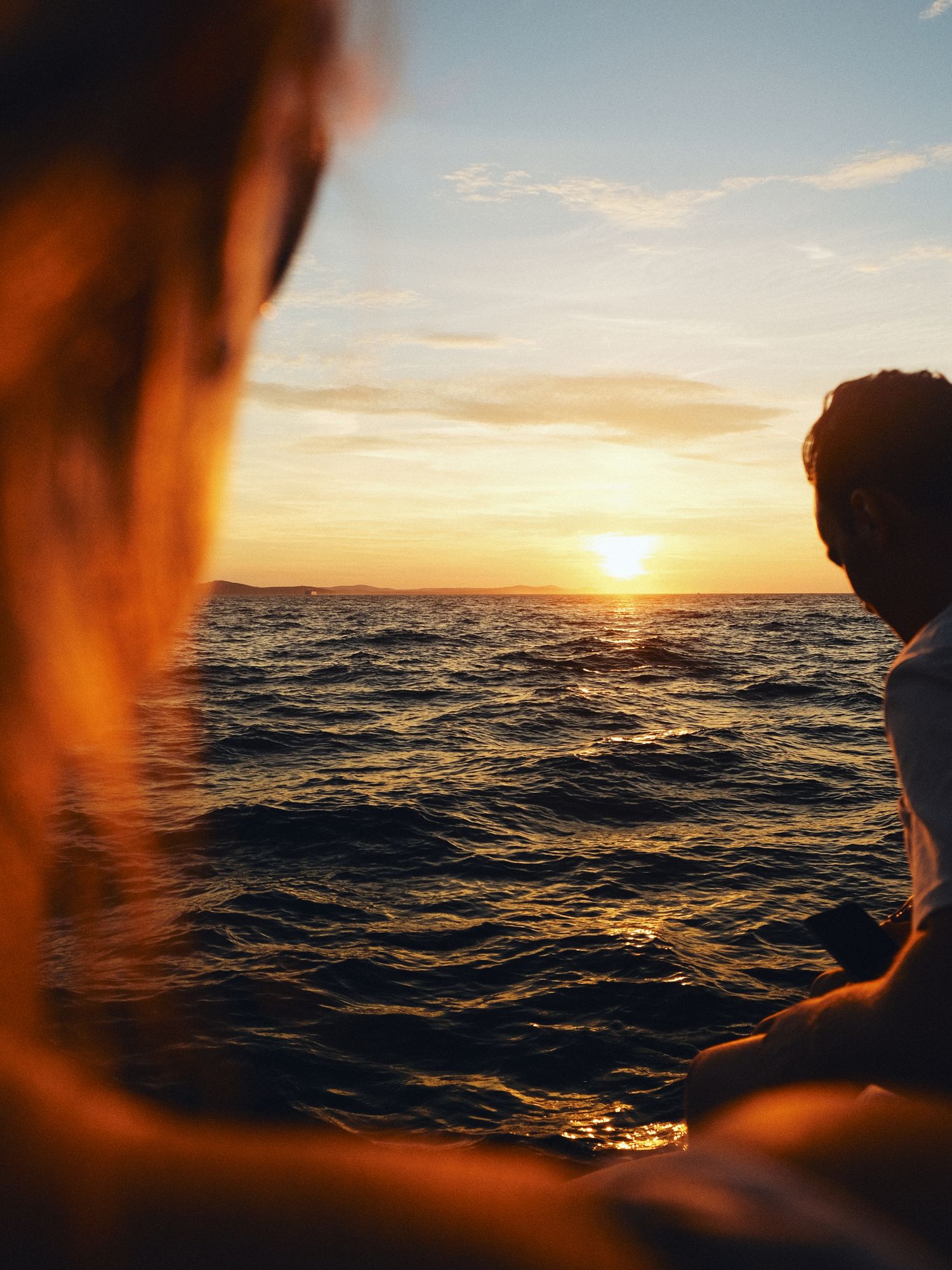 Person wearing black wetsuit relaxing on a white boat deck with coastal scenery and blue ocean waters in background.