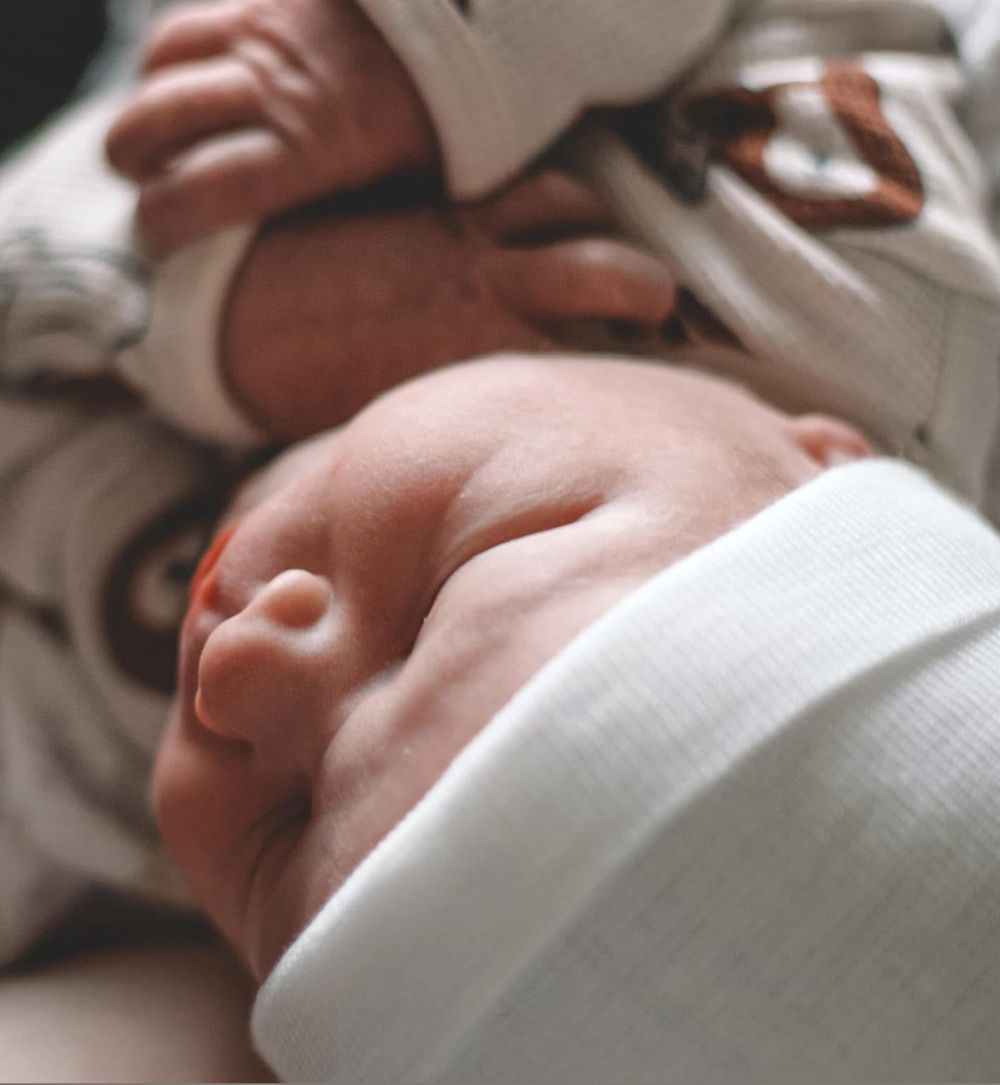 Close-up of a sleeping newborn baby in a white hat and patterned onesie, cradled in loving arms.