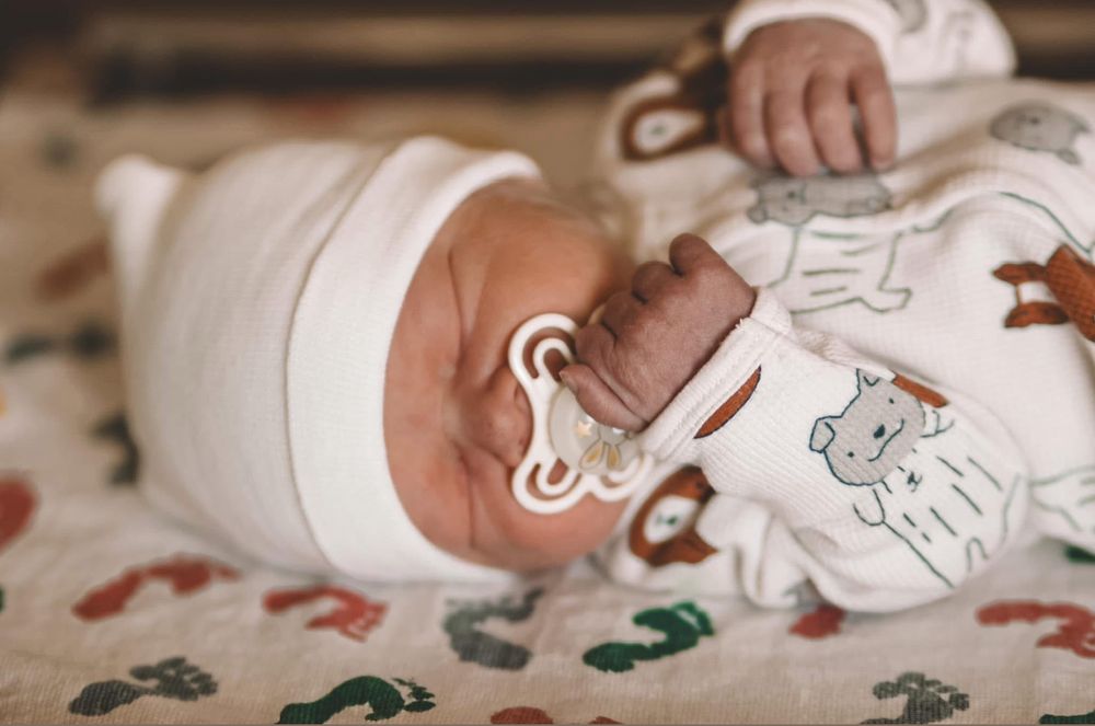Peaceful newborn baby with a pacifier lying on a colorful animal print blanket in a white outfit.